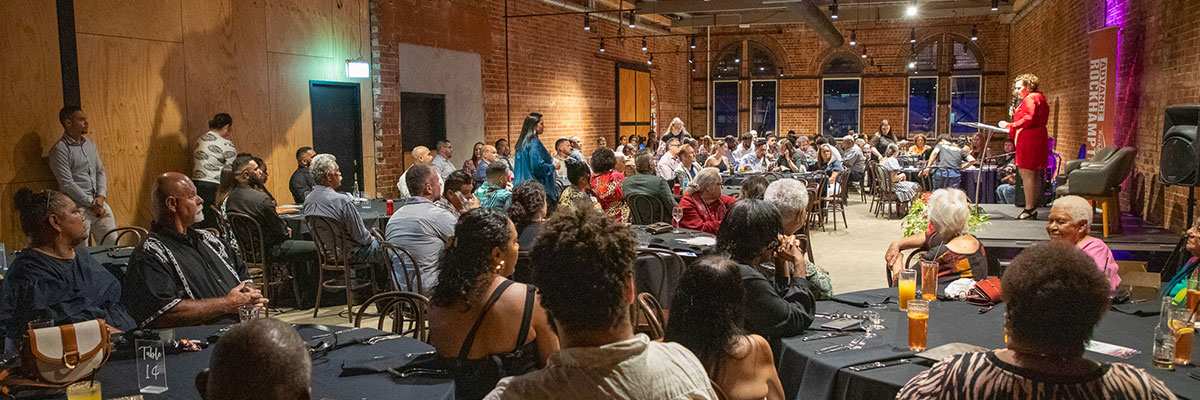 People are sitting at tables around a brick room listening to a presenter speaking on a stage