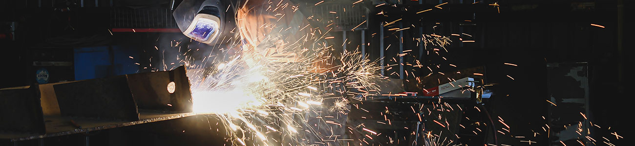 A man is wearing protective gear and using a welder which is sending sparks across a dark workshop