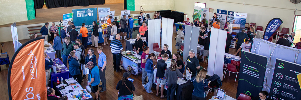 Students and students are walking past rows of exhibitors at a careers expo inside a town hall