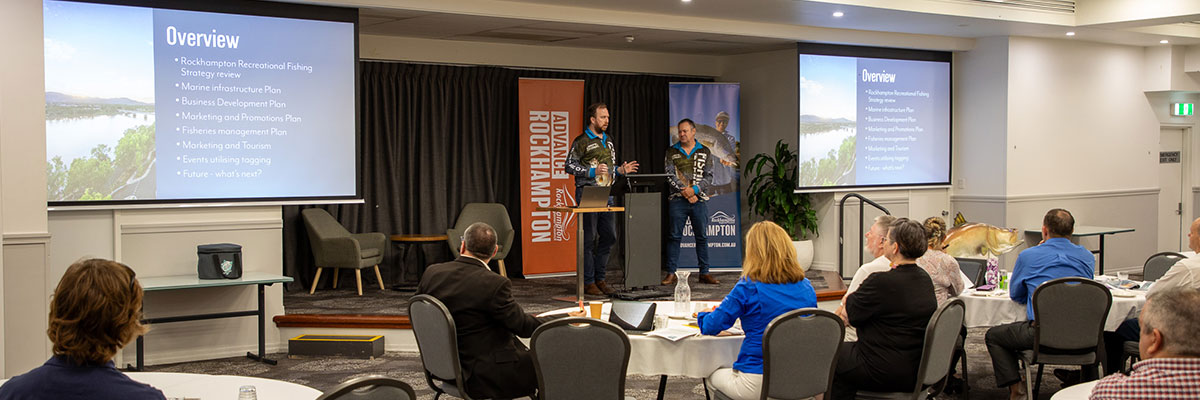 People are sitting at tables listening to two presenters on stage wearing fishing shirts